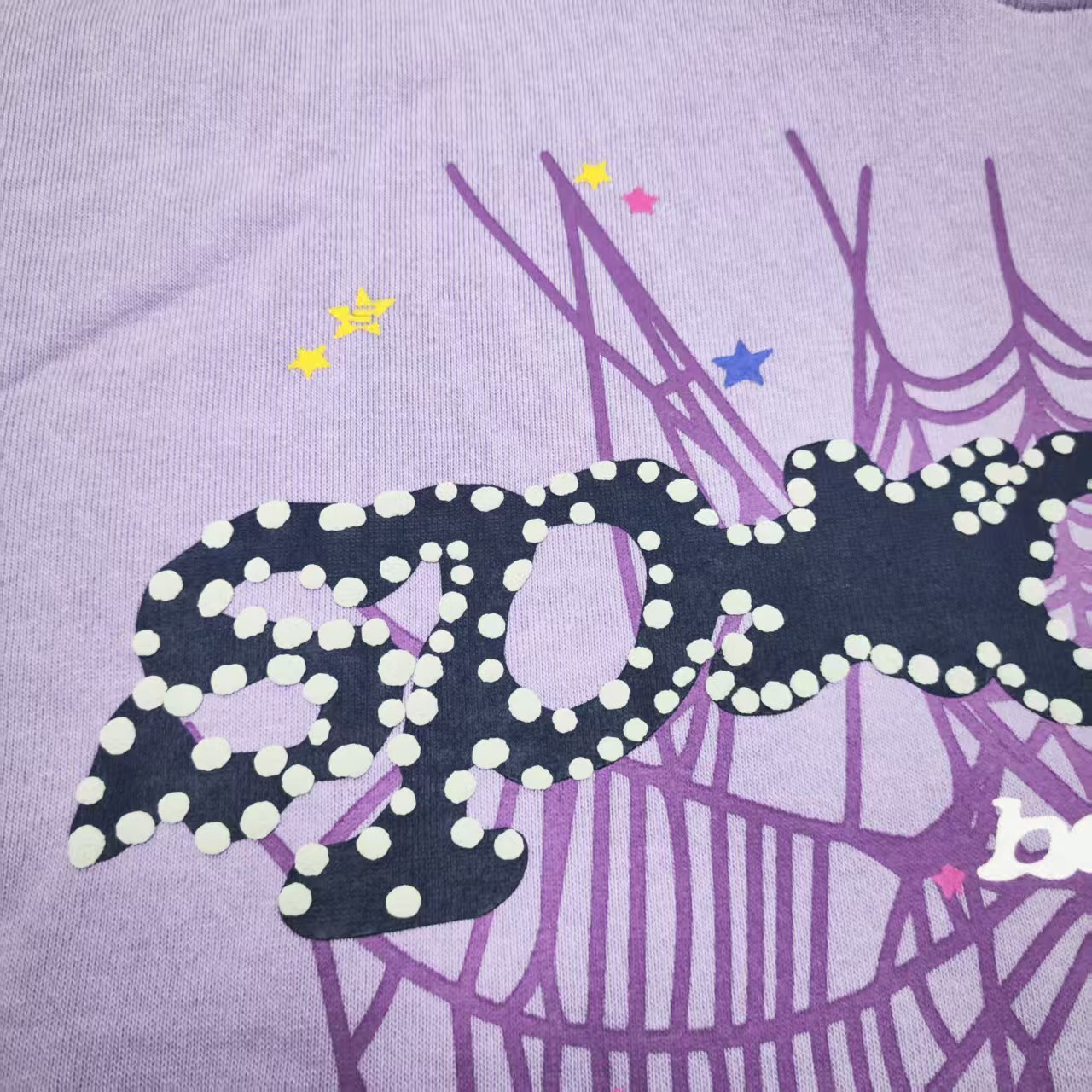 Full puff-print artwork on Sp5der Acai Hoodie Purple with black logo, tonal web and colorful stars
Close-up of raised black Sp5der lettering with dotted trim on Acai Purple Hoodie
Side angle showing the three-dimensional puff-print texture on Sp5der Acai Hoodie Purple