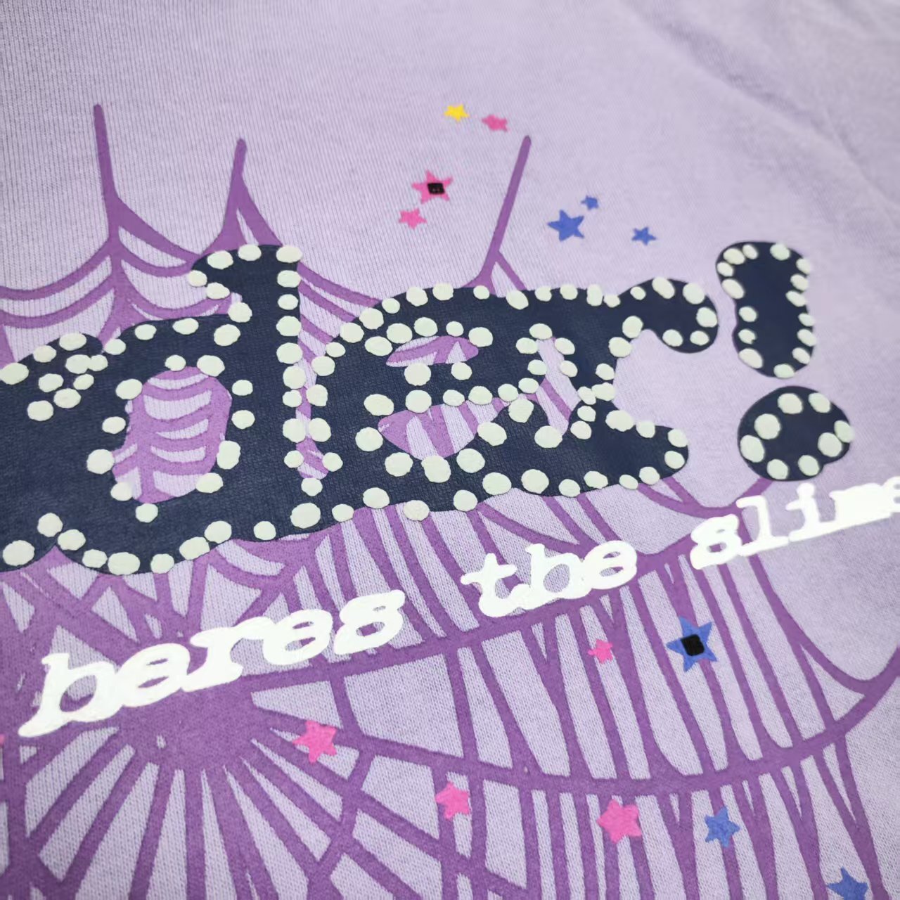 Full puff-print artwork on Sp5der Acai Hoodie Purple with black logo, tonal web and colorful stars
Close-up of raised black Sp5der lettering with dotted trim on Acai Purple Hoodie
Side angle showing the three-dimensional puff-print texture on Sp5der Acai Hoodie Purple