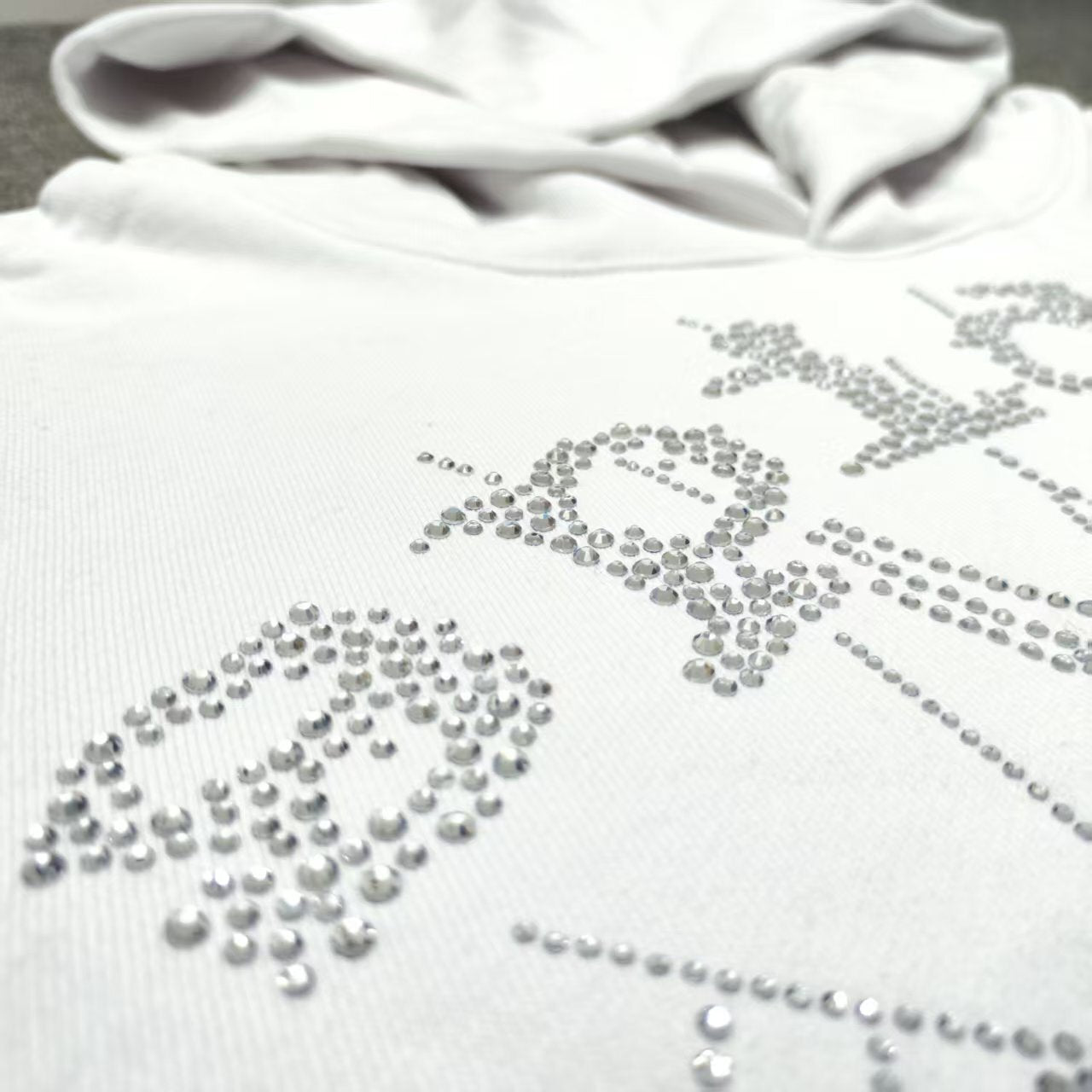 Full rhinestone artwork on Sp5der OG Logo Hoodie White with crystal lettering and spider web
Close-up of silver rhinestone Sp5der lettering and crystal web details on the white hoodie
Side angle showing the raised three-dimensional rhinestone texture on Sp5der White Hoodie