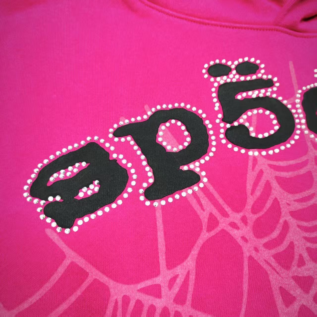 Full front artwork on Sp5der Legacy Hoodie Pink White with puff lettering, rhinestones and spider web
Close-up of raised black SP5DER puff-print lettering on the pink Legacy Hoodie
Close-up of rhinestone trim surrounding the black SP5DER lettering on the pink Legacy Hoodie
Side angle showing raised puff lettering and three-dimensional rhinestone details on Sp5der Legacy Hoodie