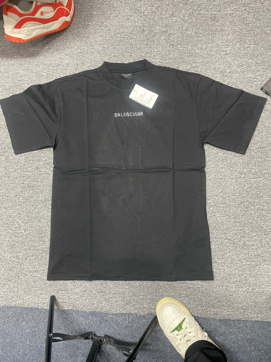 Black crew‑neck short‑sleeve t‑shirt front flat‑lay, subtle small faded Balenciaga logo on chest, paper hang‑tag attached at collar, garment laid on textured floor