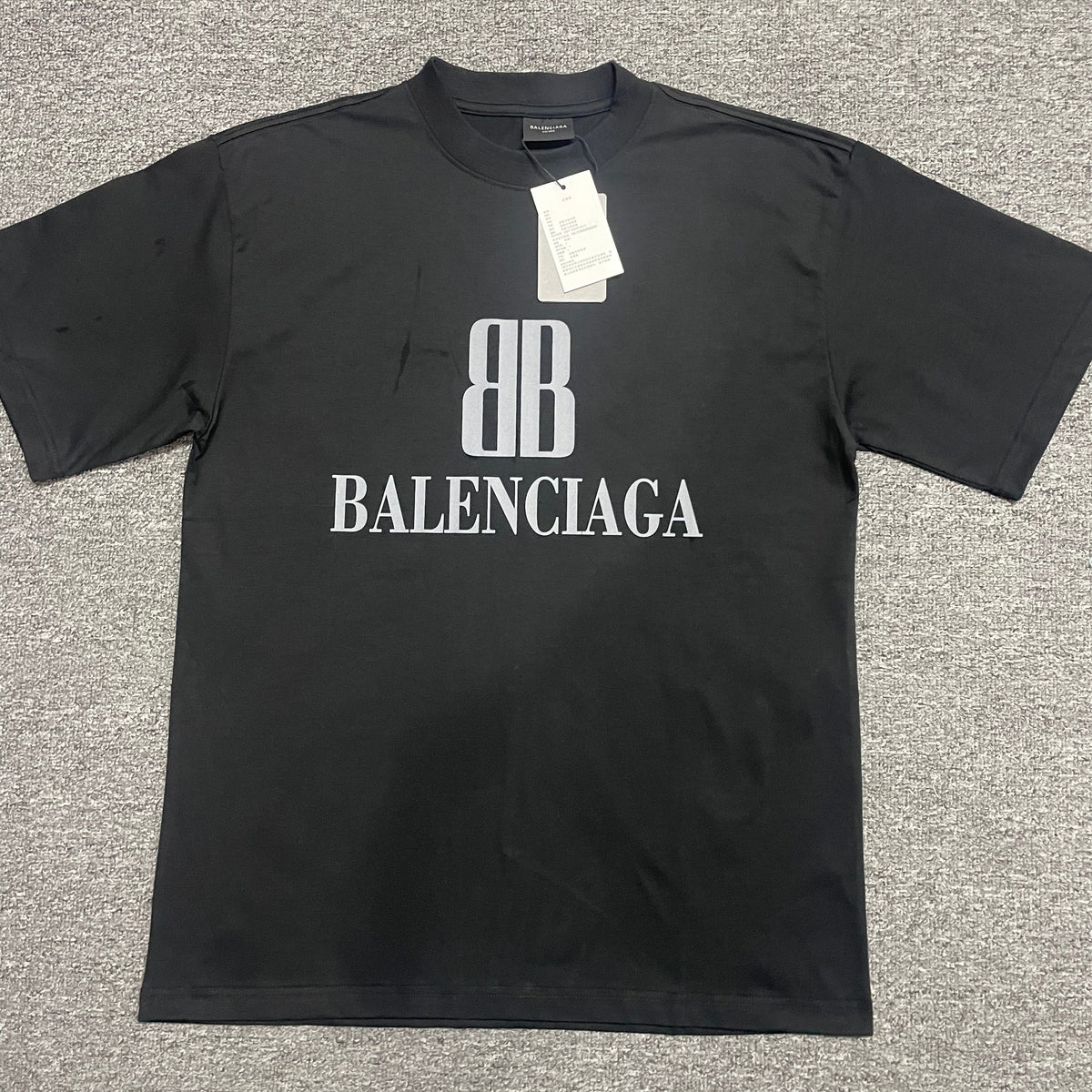 Black crew‑neck short‑sleeve t‑shirt front flat‑lay, stacked BB monogram and BALENCIAGA text print on chest, dual paper hang tags attached to collar