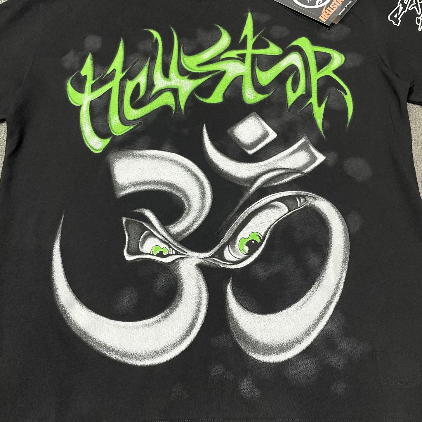 Full green and silver Om graphic on the front of the white Hellstar Reach Your Inner Peace T-Shirt
Bright green Hellstar logo printed above the Om symbol on the Reach Your Inner Peace T-Shirt
Oversized silver Om symbol with expressive eyes on the Hellstar Reach Your Inner Peace T-Shirt
Green illustrated eye details inside the Om symbol on the Hellstar Reach Your Inner Peace T-Shirt