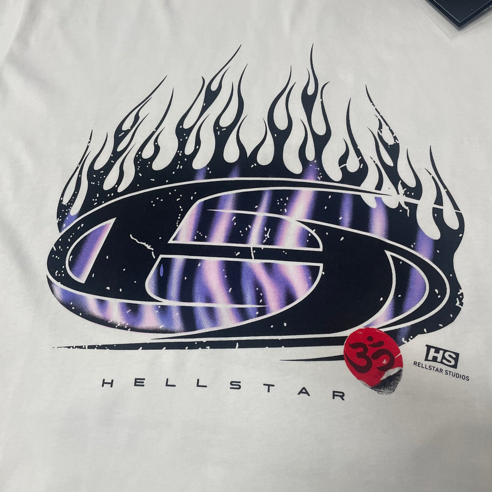 Full oval H logo and blue-purple flame graphic on Hellstar white T-shirt
Close-up of the oval H logo and blue flame print on Hellstar white T-shirt
Detailed black outlines and blue-purple flame artwork on Hellstar H Blue Flame Tee
Blue and purple flame print detail on Hellstar H Blue Flame white T-shirt
Oval H logo graphic detail on Hellstar H Blue Flame T-shirt
Red Om symbol graphic detail on Hellstar H Blue Flame white T-shirt