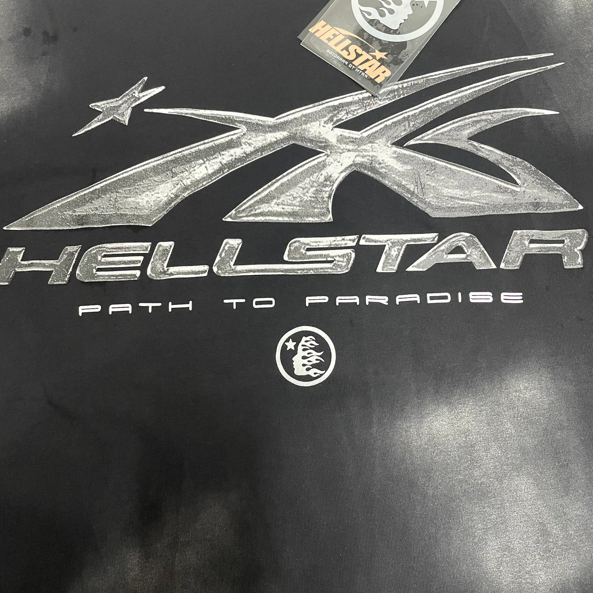 Full metallic silver Hellstar logo graphic on washed black T-shirt
Detailed metallic silver graphic print on Hellstar Chrome Logo T-shirt
Close-up of the metallic silver Hellstar logo surface
Metallic chrome-effect print texture on Hellstar black graphic T-shirt
Path to Paradise lettering detail below the Hellstar chrome logo
Hellstar shooting-star logo detail on Chrome Logo black T-shirt
Distressed black and grey wash effect across Hellstar Chrome Logo T-shirt