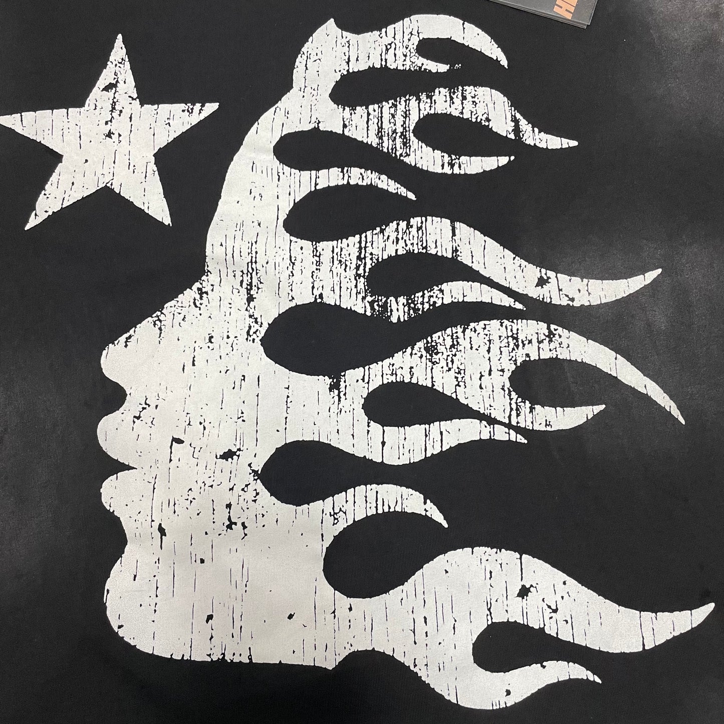 Full oversized Hellstar Flame Face and star graphic on washed black T-shirt
Close-up of white Hellstar Flame Face graphic on black T-shirt
Detailed view of distressed Hellstar Flame Face print texture
Close-up of white star graphic beside the Hellstar Flame Face design