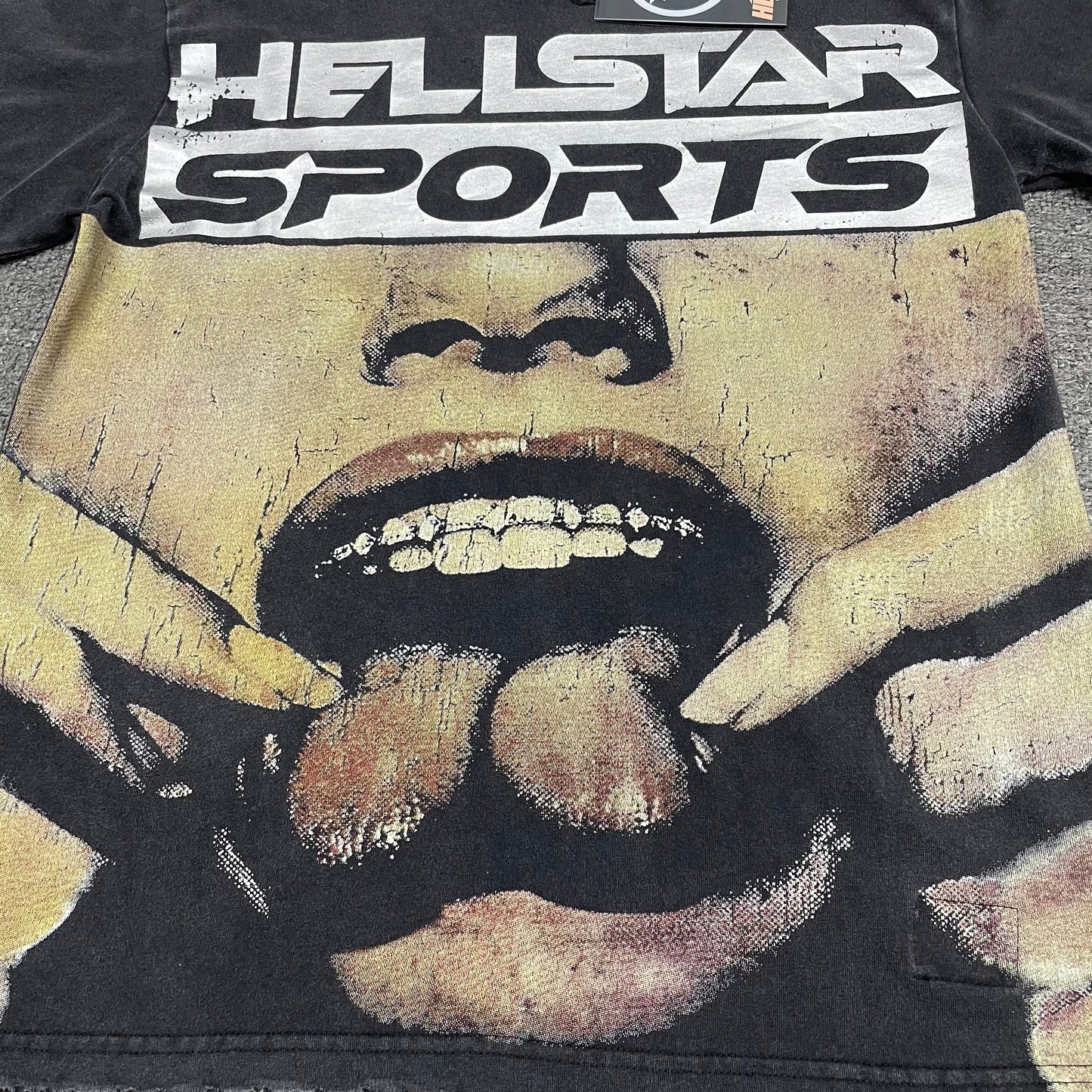 Oversized Hellstar Sports logo and mouth graphic on washed black T-shirt
Distressed Hellstar Sports lettering above a large face and mouth graphic
Close-up of white Hellstar Sports logo print on washed black T-shirt
Close-up of oversized face artwork on Hellstar Sports graphic T-shirt
Close-up of large mouth and tongue graphic on Hellstar Sports T-shirt
Detailed view of teeth lips and tongue artwork on Hellstar Sports T-shirt
Close-up of hand and finger details surrounding the mouth graphic
