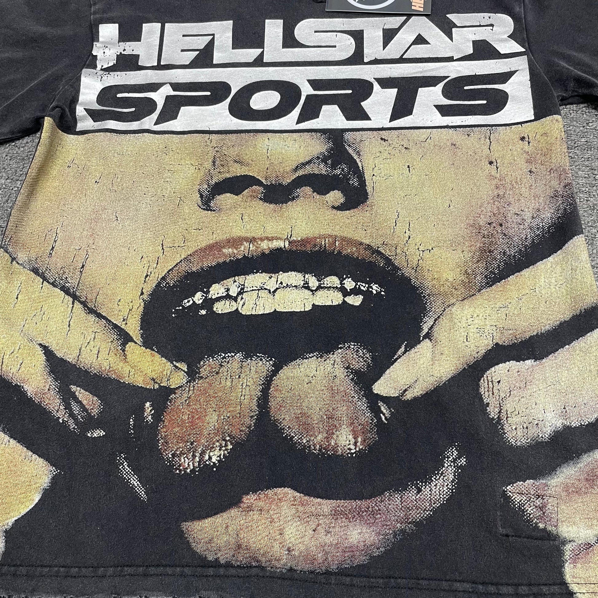 Oversized Hellstar Sports logo and mouth graphic on washed black T-shirt
Distressed Hellstar Sports lettering above a large face and mouth graphic
Close-up of white Hellstar Sports logo print on washed black T-shirt
Close-up of oversized face artwork on Hellstar Sports graphic T-shirt
Close-up of large mouth and tongue graphic on Hellstar Sports T-shirt
Detailed view of teeth lips and tongue artwork on Hellstar Sports T-shirt
Close-up of hand and finger details surrounding the mouth graphic
