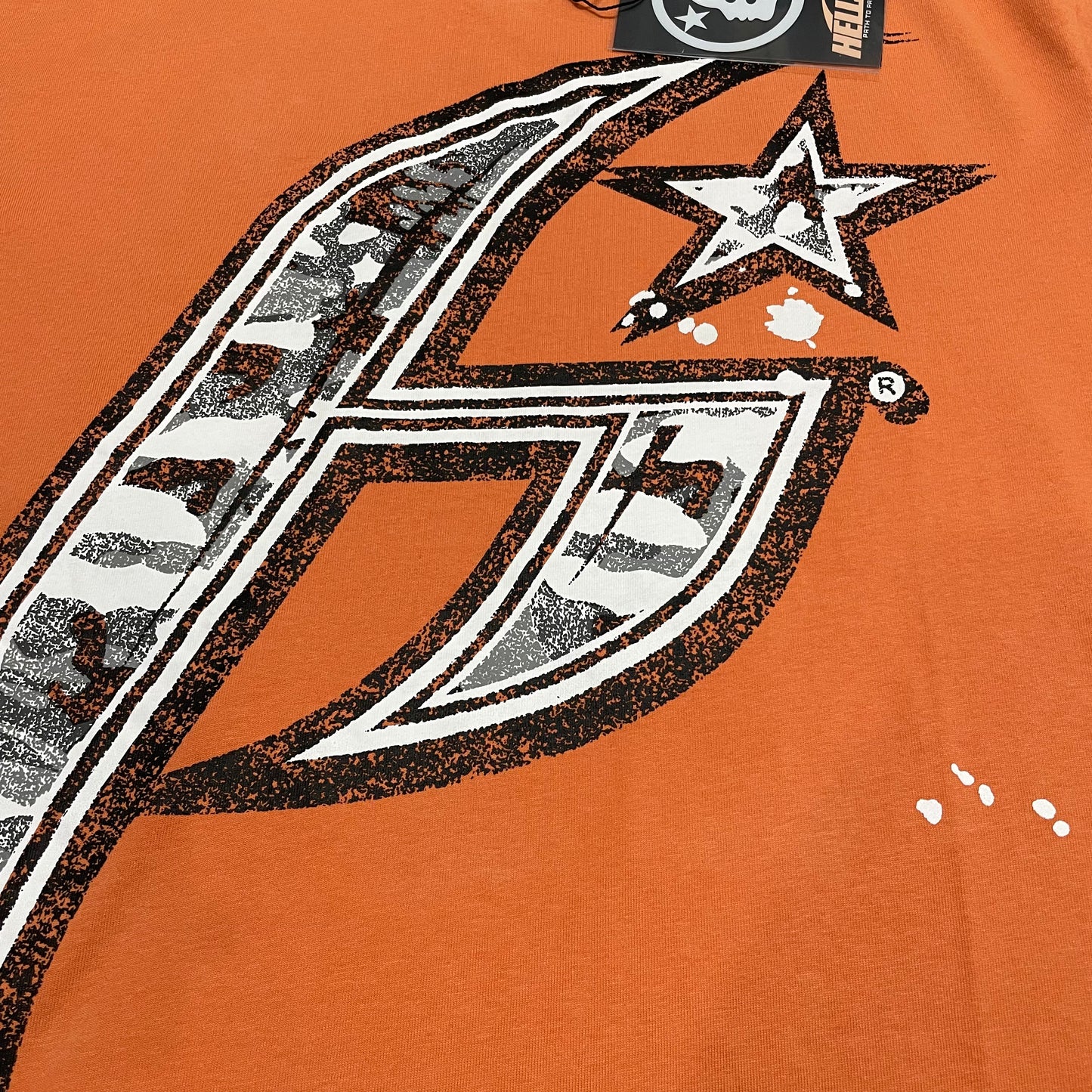 Oversized Hellstar H Sharp star logo on orange T-shirt
Large black white and silver H Sharp logo with distressed texture
Close-up of oversized Hellstar H Sharp star logo print
Detailed view of distressed black white and silver front graphic
White paint-splatter graphic details on Hellstar orange T-shirt
Close-up of distressed print texture on Hellstar H Sharp T-shirt