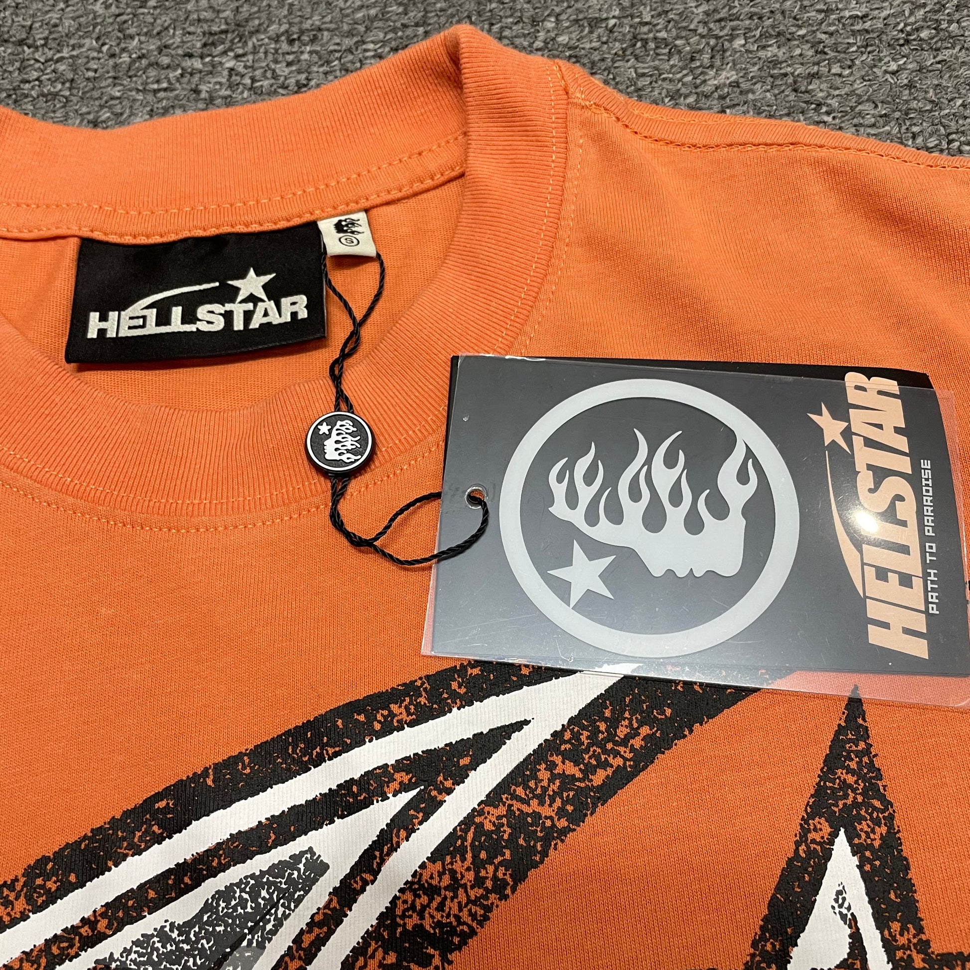 Close-up of ribbed crew neck and neckline stitching on Hellstar T-shirt
Hellstar branded neck label inside H Sharp orange T-shirt
Size label inside Hellstar H Sharp graphic T-shirt
Hellstar branded hang tag attached to H Sharp orange T-shirt