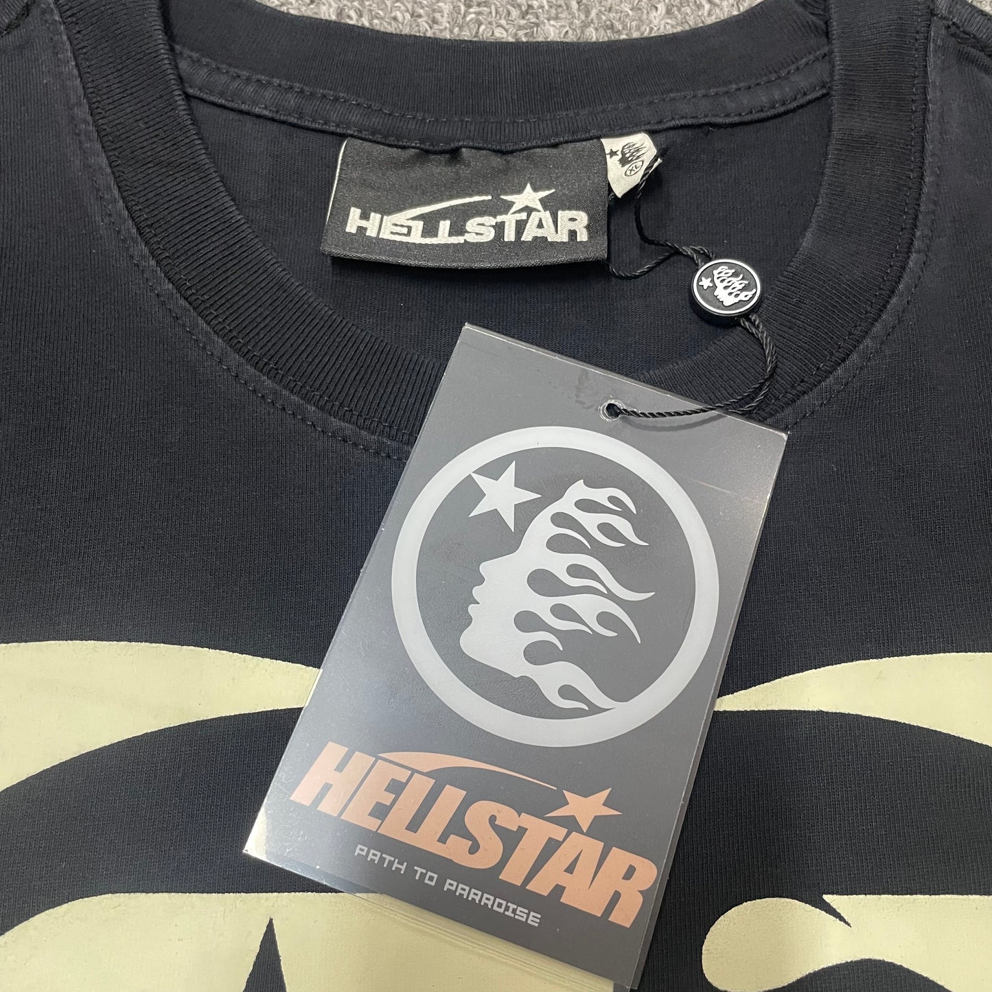 Close-up of ribbed crew neck and neckline stitching on Hellstar T-shirt
Hellstar branded neck label inside Fist Bump graphic T-shirt
Size label inside Hellstar Fist Bump washed black T-shirt
Hellstar branded hang tag attached to Fist Bump graphic T-shirt
Close-up of Hellstar flame-face hang tag and attachment detail