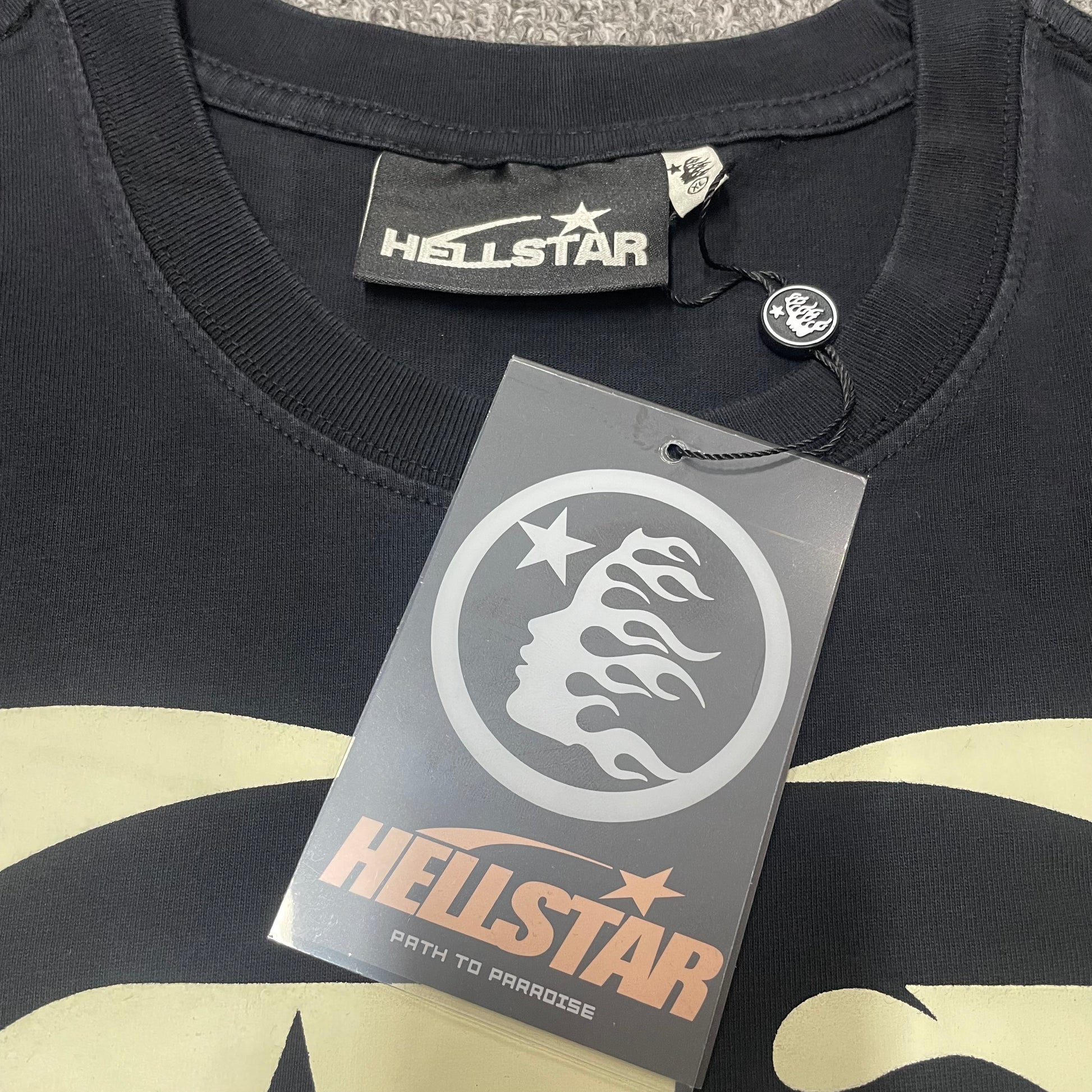 Close-up of ribbed crew neck and neckline stitching on Hellstar T-shirt
Hellstar branded neck label inside Fist Bump graphic T-shirt
Size label inside Hellstar Fist Bump washed black T-shirt
Hellstar branded hang tag attached to Fist Bump graphic T-shirt
Close-up of Hellstar flame-face hang tag and attachment detail
