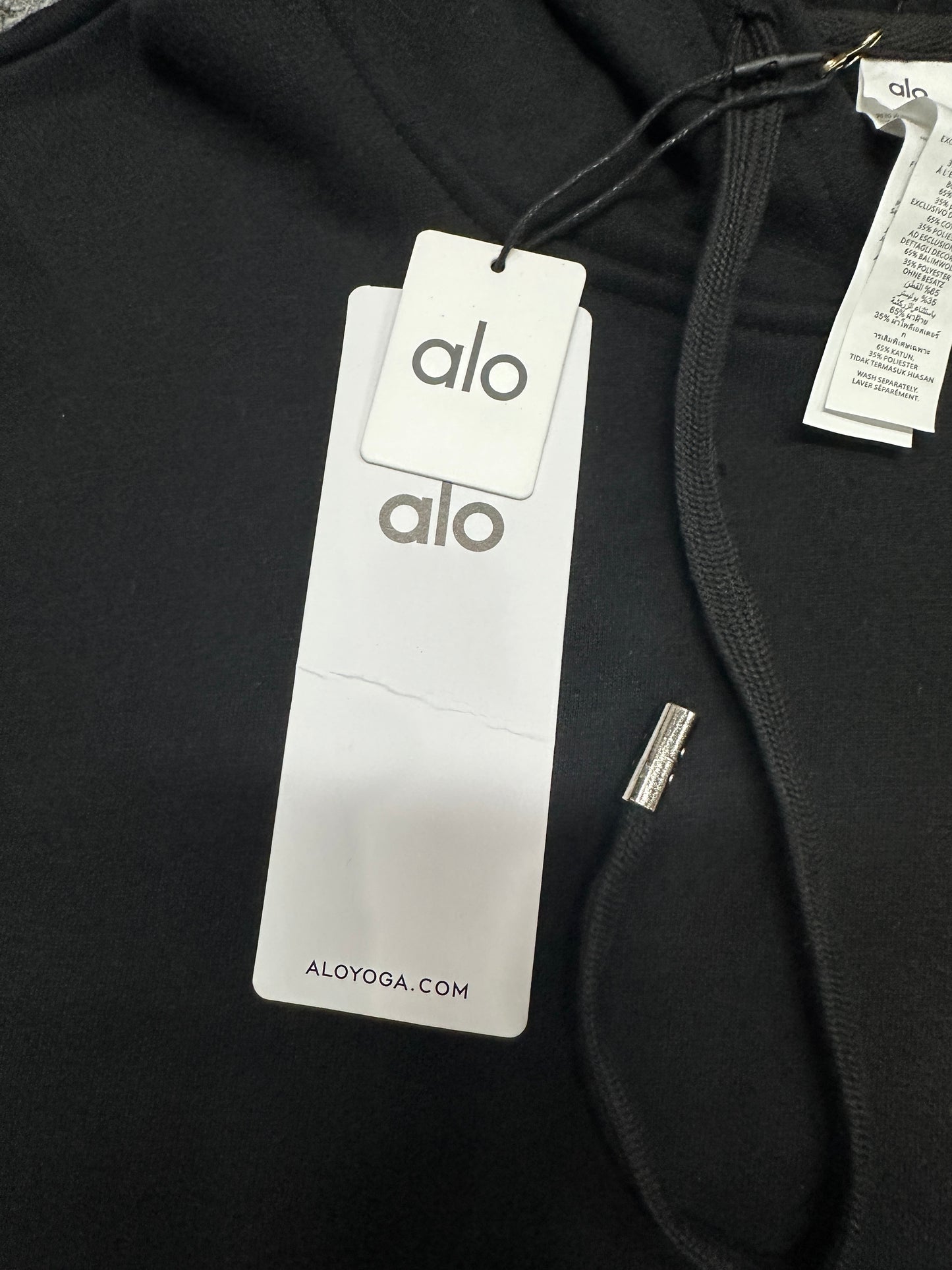 ALO Accolade Black hoodie neck label and size label close-up
ALO Accolade Black hoodie with branded retail hangtags
ALO Accolade Black hoodie care label and fabric information detail