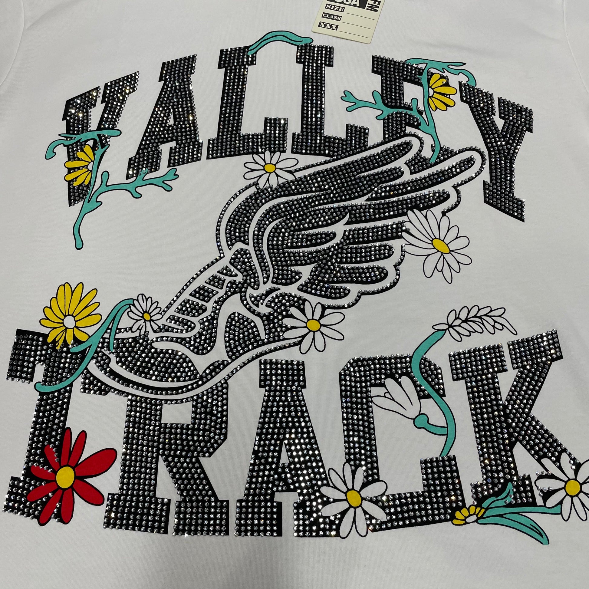Vale Forever Valley Track Tee full winged shoe and floral front graphic
Vale Forever Valley Track Tee winged track-shoe and floral artwork
Vale Forever Valley Track Tee detailed floral and wing graphic close-up
Vale Forever Valley Track Tee crystal Valley lettering detail
Vale Forever Valley Track Tee crystal Track lettering detail
Vale Forever Valley Track Tee black crystal embellishment close-up
Vale Forever Valley Track Tee winged running shoe graphic detail
